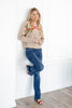 A woman modeling a beige knit sweater with green leaf embroidery and a red bow motif on the chest, paired with blue jeans and red heel sandals.