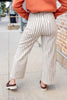 A woman shown from the back wearing cream wide-leg pinstripe pants with thin dark vertical stripes, an orange sweatshirt, and brown slip-on shoes on a sidewalk next to a brick wall.