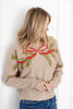 A woman wearing a beige knit sweater with a red bow and green pine branch pattern, paired with blue jeans.