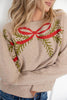 A woman wearing a beige knit sweater with a red bow and green pine branch pattern across the chest, paired with blue jeans.
