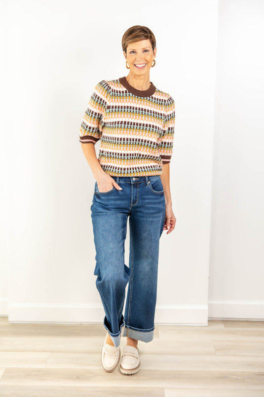 A woman modeling a short-sleeve knit top with a multicolor geometric pattern in beige, brown, pink, and sage, paired with blue jeans and white slip-on loafers.