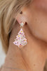 A close-up of a woman ear wearing a dangling Christmas tree earring. The earring is light pink with rhinestone embellishments in pink, yellow, orange, and silver, topped with a small silver star.