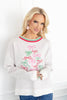 A woman wearing a white crewneck sweater with a colorful embroidered Christmas tree made of pink and green bows.