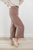 A woman wearing brown wide-leg pants with a white polka dot pattern and ruffled hems, styled with a cream knit sweater and cream slip-on shoes against a white wall and light wood floor.