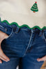 Close-up of a woman wearing a cream ribbed knit sweater with a green scalloped hem and a small embroidered Christmas tree motif, paired with blue jeans.
