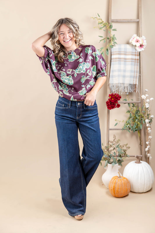 A woman modeling a short-sleeve blouse with a green and teal floral pattern on a deep purple background, paired with dark blue wide-leg jeans, in a styled studio setting with pumpkins, a ladder, and a plaid throw.