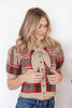A woman wearing a short-sleeve knit cardigan with a red, green, and beige plaid pattern and a knotted tie neckline, paired with blue jeans.