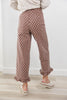 A woman wearing brown wide-leg pants with a white polka dot pattern and ruffled hems, paired with a cream knit sweater and white slide sandals, standing on a light wood floor against a white wall.