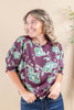 A woman wearing a purple short-sleeve blouse with a green and white floral pattern, paired with blue jeans, posing against a neutral beige background.