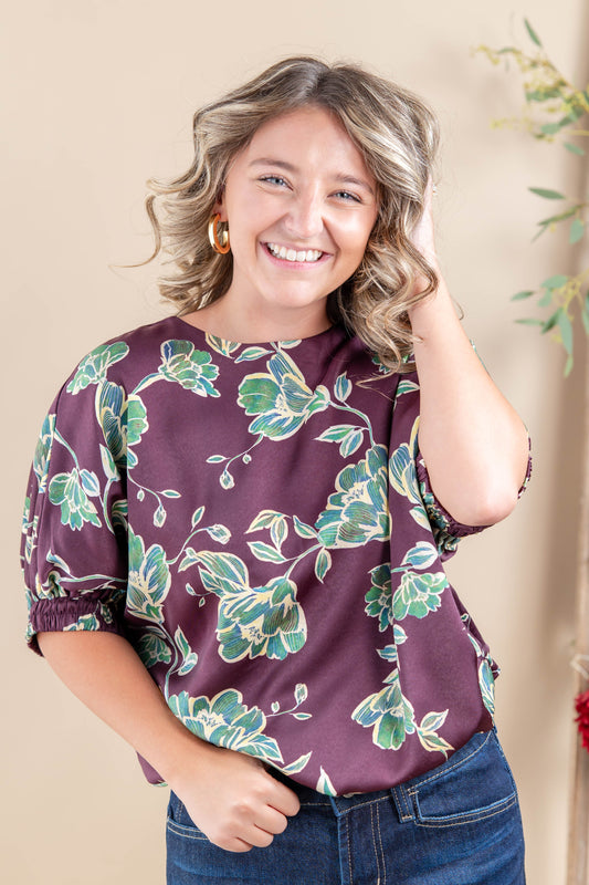 A smiling woman with blonde-highlighted hair wearing a purple short-sleeve blouse with a green and beige floral pattern, paired with blue jeans, standing against a warm beige background.