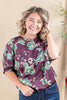 A smiling woman with blonde-highlighted hair wearing a purple short-sleeve blouse with a green and beige floral pattern, paired with blue jeans, standing against a warm beige background.