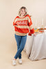 A woman wearing a festive orange and beige striped sweater with 'COBBLE' written across the chest, paired with blue jeans and white shoes, posing next to a decorated table with candles.
