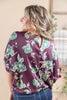 A woman shown from the back wearing a purple satin blouse with a green and white floral pattern, paired with blue jeans in an indoor setting.