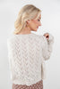 A blonde woman shown from the back wearing a cream pointelle-knit sweater with a textured chevron pattern, paired with brown polka-dot pants, against a light gray studio background.