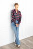 A woman stands wearing a navy, red and black plaid button-up shirt with a ruffle collar, light blue jeans, and beige boots.
