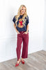 A woman stands wearing a navy blue knit top with a sitting dog graphic, burgundy cropped pants, and red pointed flats.