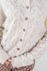 Close-up of a person wearing a cream-colored open-knit pointelle cardigan sweater with brown buttons, paired with brown polka-dot pants.
