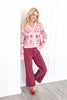 A woman stands wearing a pink holiday knit cardigan with red and green festive patterns, paired with burgundy wide-leg pants and red pointed flats.