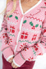 A close-up of a woman wearing a pink knit cardigan with a festive Christmas pattern featuring red gifts, green holly, and white zigzags and borders.