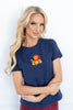 A woman wearing a navy blue short-sleeve t-shirt with a small needlework graphic of a teddy bear holding a red Santa hat and a candy cane on the left chest.