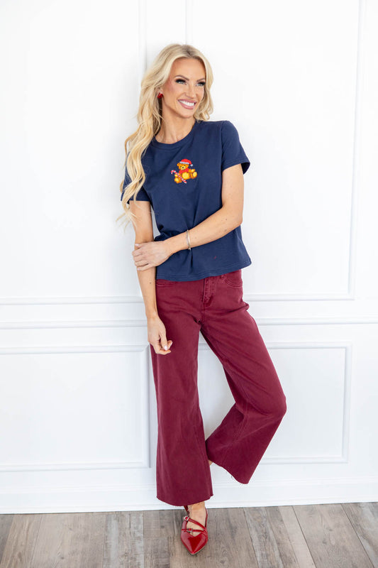 A woman stands wearing a navy blue short sleeve t-shirt with a small red and yellow bear needlework graphic on the left chest, paired with burgundy wide-leg pants and red pointed flats.
