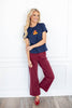 A woman stands wearing a navy blue short sleeve t-shirt with a small red and yellow bear needlework graphic on the left chest, paired with burgundy wide-leg pants and red pointed flats.