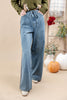 A person wearing medium wash wide-leg denim jeans with a drawstring waist, styled with an olive green top and white shoes in a warm beige studio setting with pumpkins and decorative plants.