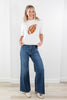 A woman stands in a bright indoor room wearing a cream knit sweater with a brown football graphic on the front, paired with wide-leg blue jeans and beige shoes.