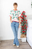A woman wearing a short-sleeve knit sweater with a Christmas holly and berry pattern in green, red, and white paired with denim.