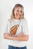 A smiling blonde woman wearing a cream knit sweater with a brown and white football graphic on the front, paired with blue jeans, against a plain light gray studio background.