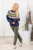 A woman modeling a navy blue knit sweater with a colorful Fair Isle yoke pattern in orange, green, yellow, and white, paired with olive green pants and beige boots.