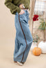 A person wearing wide-leg light blue denim jeans with a dark brown side stripe detail, styled with an olive green cardigan and beige shoes in a warm beige studio setting with pumpkins and autumn decor.