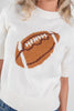 A close-up of a woman wearing a cream knit sweater with a large brown and white football graphic on the front.