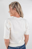 A woman shown from the back wearing a cream-colored knit sweater with short puff sleeves and ribbed trim, paired with blue jeans, against a plain light gray studio background.