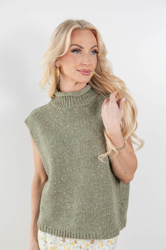 A blonde woman wearing a sleeveless ribbed knit mock neck sweater vest in a heathered sage green color, paired with a floral skirt, against a plain light gray background.
