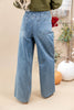 A person shown from the back wearing light blue wide-leg denim jeans, with an olive green top partially visible. The setting is a styled indoor scene with a beige backdrop, decorative pumpkins, and plants.