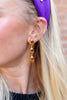 A close-up of a woman's ear wearing a dangling triple-stacked candy corn earring. The earring features amber-orange and yellow textured beads with small clear rhinestone accents at the top and bottom, set in gold-toned metal.