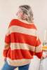 A woman wearing a chunky knit sweater with wide horizontal stripes in orange and cream, shown from a back-three-quarter view in an indoor setting.