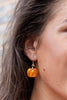A close-up of a woman's ear wearing a dangling pumpkin-shaped earring with orange and yellow stripes and small red rhinestone accents.