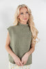 A blonde woman wearing a sleeveless ribbed knit mock neck sweater in a heathered sage green color, paired with a white and yellow floral skirt, against a plain light gray background.