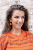 A smiling young woman with long brown hair wears small orange pumpkin-shaped drop earrings and a matching orange pumpkin pendant necklace, paired with an orange and brown striped sweater, standing against a light brick wall.
