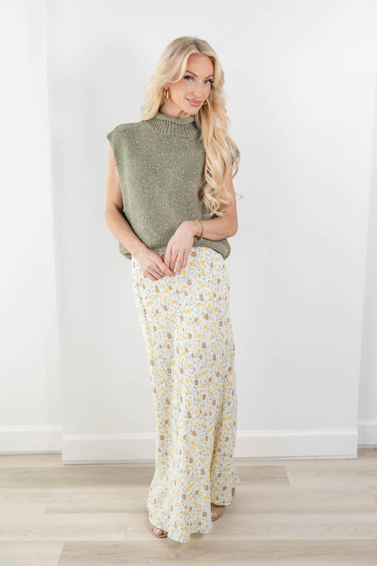 A woman stands in a bright indoor room wearing a sleeveless olive green knit top and a cream midi skirt with a small yellow and blue floral print.