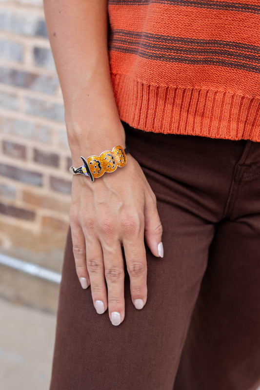 A close-up of a person's hand wearing a chunky orange and black stacked jack-o-lantern cuff bracelet, paired with an orange striped sweater and brown pants.