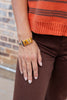 A close-up of a person's hand wearing a chunky orange and black stacked jack-o-lantern cuff bracelet, paired with an orange striped sweater and brown pants.