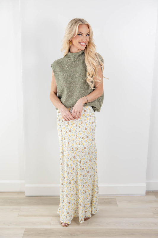 A woman modeling a sleeveless sage green mock neck knit top and a long cream maxi skirt with a small yellow and green floral print, standing in a bright room with light wood floors.