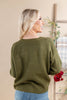 A woman with blonde hair is shown from the back wearing an olive green knit sweater and blue jeans in a warm indoor setting.