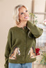 A smiling blonde woman wearing an olive green knit cardigan with embroidered horse designs on the front, paired with blue jeans in a softly decorated indoor setting.