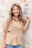 A smiling woman with wavy light brown hair stands against a light gray brick wall, wearing a tan and white gingham short-sleeve button-up shirt and blue jeans.