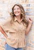 A smiling woman with wavy light brown hair stands against a brick wall, wearing a tailored tan and white gingham short-sleeve button-up shirt, blue skinny jeans.