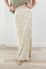 A person wearing a long cream maxi skirt with a small yellow and brown floral print, paired with a green top and beige sandals, standing on a light wood floor.